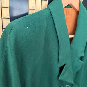 Peut inclure: Un blazer vert fonc&eacute; avec un revers crant&eacute; et quatre boutons fonc&eacute;s. Le blazer est suspendu &agrave; un cintre en bois, r&eacute;v&eacute;lant une doublure orange. Le tissu semble &ecirc;tre de poids moyen.