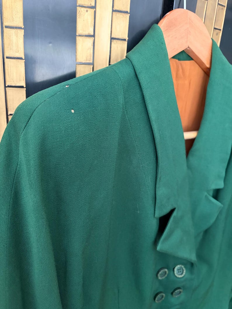 Peut inclure: Un blazer vert fonc&eacute; avec un revers crant&eacute; et quatre boutons fonc&eacute;s. Le blazer est suspendu &agrave; un cintre en bois, r&eacute;v&eacute;lant une doublure orange. Le tissu semble &ecirc;tre de poids moyen.
