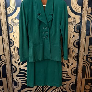 Peut inclure: Un tailleur jupe vert vintage compos&eacute; d'un blazer et d'une jupe mi-longue. Le blazer a un col crant&eacute;, quatre boutons et deux poches avant. Le costume est fait d'un tissu doux, peut-&ecirc;tre du velours, et est suspendu &agrave; un cintre en bois.