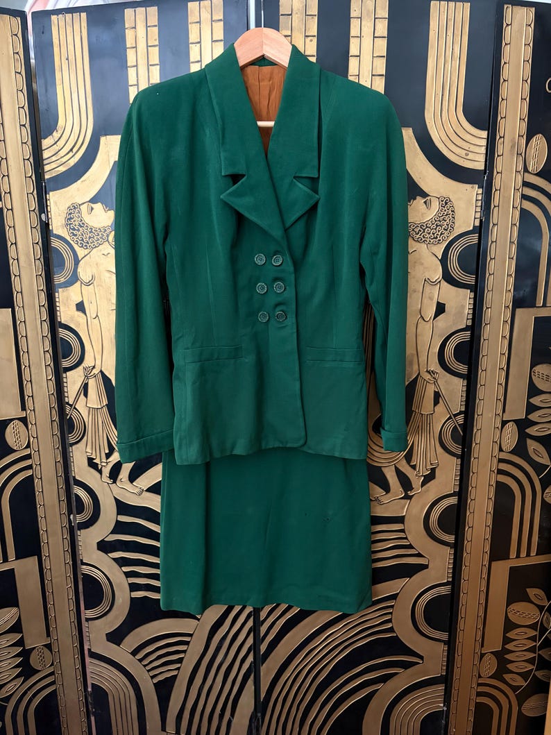 Peut inclure: Un tailleur jupe vert vintage compos&eacute; d'un blazer et d'une jupe mi-longue. Le blazer a un col crant&eacute;, quatre boutons et deux poches avant. Le costume est fait d'un tissu doux, peut-&ecirc;tre du velours, et est suspendu &agrave; un cintre en bois.