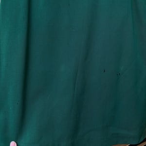 Peut inclure: Gros plan sur un tissu vert fonc&eacute;, possiblement un v&ecirc;tement ou un textile. Le tissu a une texture lisse avec de subtiles variations de couleur et quelques petites imperfections. Le bord inf&eacute;rieur du tissu est visible, et une main est partiellement visible.