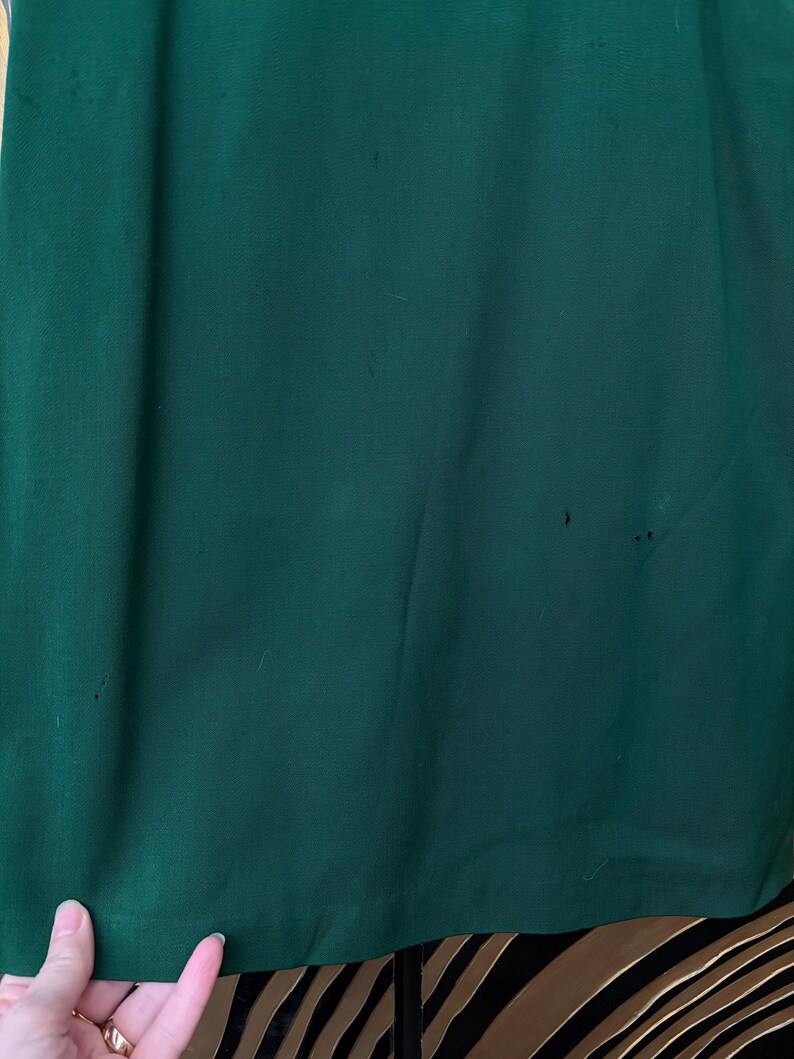 Peut inclure: Gros plan sur un tissu vert fonc&eacute;, possiblement un v&ecirc;tement ou un textile. Le tissu a une texture lisse avec de subtiles variations de couleur et quelques petites imperfections. Le bord inf&eacute;rieur du tissu est visible, et une main est partiellement visible.