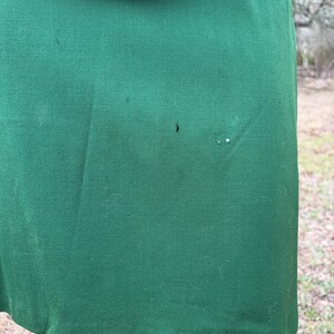Peut inclure: Gros plan sur une jupe vert fonc&eacute; et le bord d'une veste assortie. Le tissu a une texture lisse avec quelques petites imperfections. La jupe est l&eacute;g&egrave;rement incurv&eacute;e &agrave; l'ourlet. La tenue semble &ecirc;tre vintage.