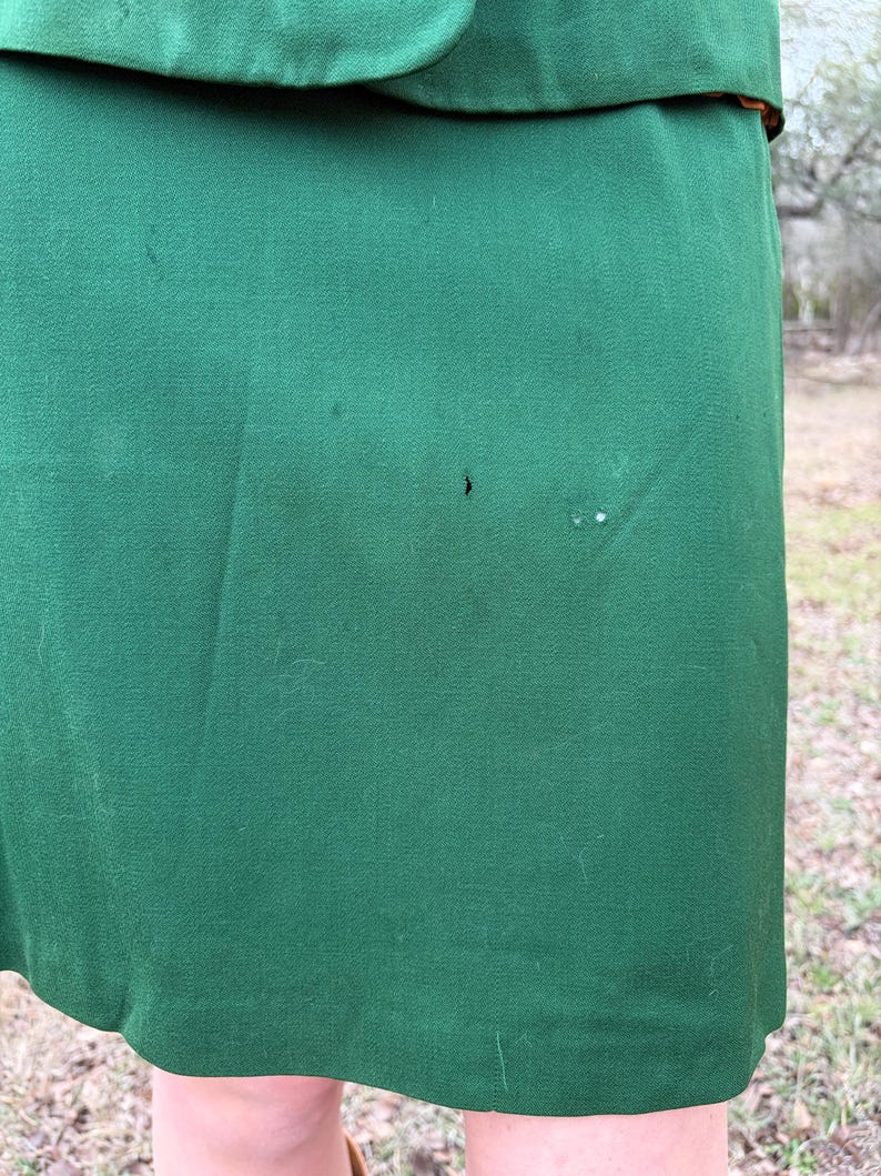 Peut inclure: Gros plan sur une jupe vert fonc&eacute; et le bord d'une veste assortie. Le tissu a une texture lisse avec quelques petites imperfections. La jupe est l&eacute;g&egrave;rement incurv&eacute;e &agrave; l'ourlet. La tenue semble &ecirc;tre vintage.