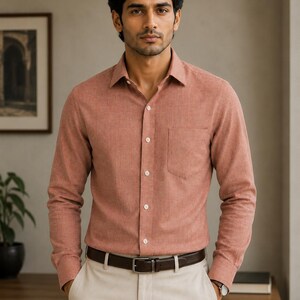 May include: A long-sleeved button-down shirt in a muted terracotta orange color with a subtle textured finish, featuring a classic collar, a single chest pocket, and white buttons, paired with beige trousers and a dark brown leather belt.
