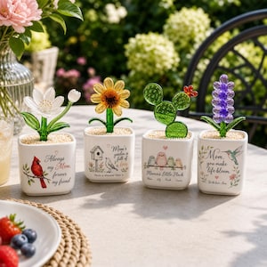 May include: Four square white ceramic planters containing glass flower and cactus sculptures. Each planter features unique printed text and illustrations, including a red cardinal, a birdhouse, small birds, and a hummingbird with floral accents.