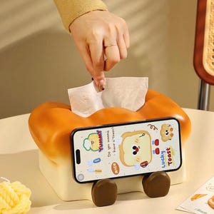 May include: A rectangular tissue box holder shaped like a loaf of bread with a golden-brown crust and a cream-colored base. The front features two brown rounded supports for holding a smartphone, which displays a cartoon toast graphic and text.