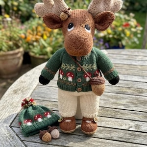 May include: A hand-knitted moose doll with brown fur, large antlers, and black eyes. It wears a green cardigan with a red and white mushroom pattern, cream-coloured trousers, and brown shoes. A small acorn bag hangs from its shoulder, and a matching hat sits nearby.