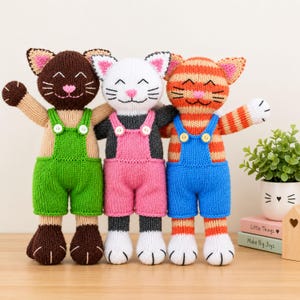 Pode incluir: Três bonecos de gato tricotados à mão lado a lado. O gato da esquerda é castanho com jardineiras verdes, o central é branco e cinzento com jardineiras cor-de-rosa, e o da direita é às riscas cor de laranja com jardineiras azuis. Rostos bordados.