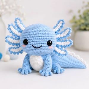 Peut inclure: Une peluche axolotl crochetée bleu clair avec un ventre blanc, de grands yeux noirs et des marques de blush roses. La créature présente des branchies externes plumeuses proéminentes en bleu et blanc, ainsi qu'une longue queue et de petits pieds.