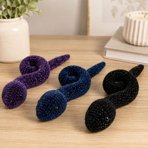 Puede incluir: Tres serpientes de ganchillo hechas con hilo brillante en morado, azul oscuro y negro. Cada serpiente presenta un cuerpo en espiral, una cabeza redondeada y pequeños ojos de cuentas negras. El material texturizado y afelpado refleja la luz.