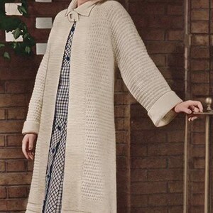 May include: A long, cream-colored knit cardigan featuring a textured pattern, a collared neckline, and a single button closure at the top. Underneath, a blue and white checkered dress with a button-down front is visible.