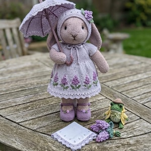 May include: A knitted rabbit doll wearing a lavender dress with floral embroidery, a matching bonnet, and shoes, holding a lavender umbrella. Beside the rabbit is a small green knitted frog wearing a hat and coat, and a square lace-trimmed cloth.
