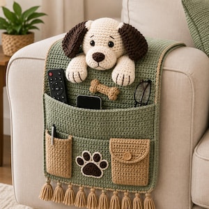 May include: A sage green crochet armchair organizer featuring a stuffed dog head with dark brown ears, a bone-shaped accent, and a paw print motif. It includes multiple pockets holding a remote, phone, glasses, and pen, finished with tan tassels.