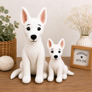 May include: Two white crochet dog figures with pointed ears, black noses, and dark eyes. The larger figure sits on the left, and the smaller figure sits on the right. Both are crafted with a textured yarn stitch, featuring pink inner ears.