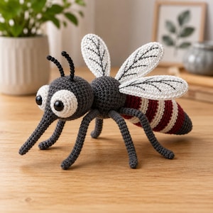 May include: A crocheted mosquito toy featuring a dark gray head and body, large white eyes with black pupils, two black antennae, and four white wings with black stitched veins. The abdomen has alternating dark red and white horizontal stripes.
