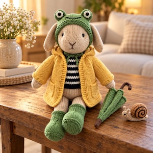 May include: A knitted stuffed rabbit toy wearing a green frog-themed hat, a yellow hooded jacket, a black and white striped sweater, and green booties. Beside the rabbit sits a small green knitted umbrella and a tiny brown knitted snail.