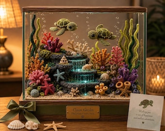 Op de afbeelding: Een glazen vitrine met een gehaakte onderwaterscène met twee zeeschildpadden, kleurrijk koraal, zeesterren en een verlichte waterval. De houten voet is voorzien van een plaatje met de tekst Ocean Garden en een groene strik.