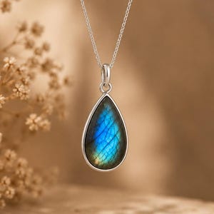 May include: A teardrop-shaped labradorite pendant set in a polished silver bezel, suspended from a delicate silver chain. The gemstone displays a vibrant blue iridescence with visible internal fractures and a smooth, rounded surface finish.