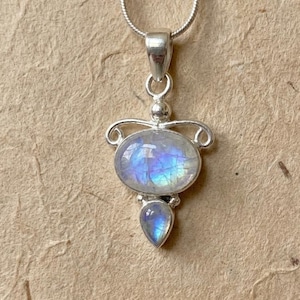 May include: A silver pendant necklace featuring a large oval moonstone and a smaller teardrop-shaped moonstone set in a decorative metal frame. The stones exhibit a blue iridescent sheen, and the pendant hangs from a thin silver chain.