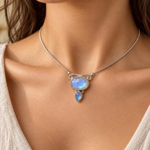 May include: A silver necklace featuring a central oval moonstone cabochon and a smaller pear-shaped moonstone drop. The stones exhibit a blue iridescent glow set within a polished silver frame, suspended from a flexible silver snake chain.