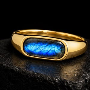 Peut inclure: Une bague en or dotée d'un anneau lisse et poli, sertie d'une pierre de labradorite ovale disposée horizontalement. La pierre présente un éclat bleu irisé et vibrant avec des fractures internes naturelles visibles en surface.
