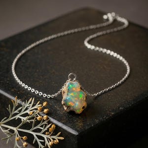 Op de afbeelding: Een zilveren ketting met een ruwe, ongepolijste opaal hanger. De steen vertoont iriserende flitsen van groen, oranje en blauw tegen een melkachtige witte basis. De hanger is bevestigd met een klein zilveren oogje aan de fijne ketting.