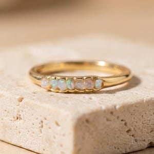 Può includere: Un anello in oro con una fila di cinque piccole opale bianche rotonde e iridescenti incastonate orizzontalmente sulla parte anteriore della fascia. L'anello in metallo ha una finitura liscia e lucida e le pietre mostrano riflessi verdi e rosa.