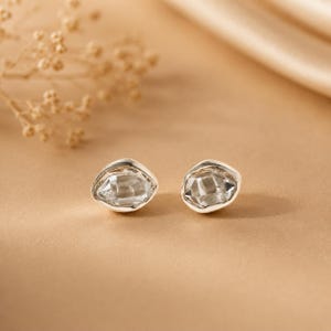 Peut inclure: Une paire de boucles d'oreilles à clous avec des cristaux transparents, facettés et de forme irrégulière, sertis dans un métal argenté poli. Les boucles présentent un bord lisse et organique autour de la pierre centrale, offrant un aspect réfléchissant.