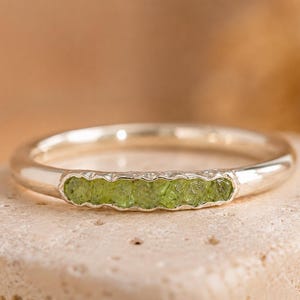 May include: A polished silver band ring featuring a horizontal row of six small, rough-cut, light green peridot gemstones set into a rectangular channel. The metal band has a smooth, reflective finish that contrasts with the textured green stones.