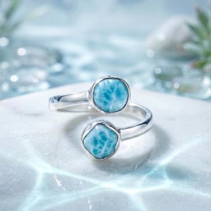 Könnte beinhalten: Ein offener Ring aus poliertem Silber mit zwei verschiedenen Larimar-Edelsteinen. Ein Stein ist kreisförmig und der andere quadratisch abgerundet, beide mit einem marmorierten himmelblauen und weißen Muster in glatten Silberfassungen.