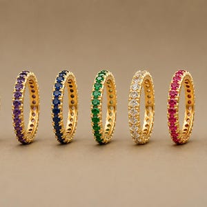 Op de afbeelding: Zeven gouden eternity-ringen opgesteld in een horizontale rij. Elke ring heeft een doorlopende band van kleine, rond geslepen edelstenen in verschillende kleuren: rood, paars, donkerblauw, groen, helder wit, roze en lichtblauw, gezet in geelgoud.