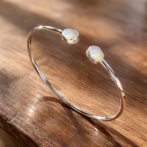 May include: A silver-toned open cuff bracelet featuring two polished, iridescent opal stones set at each end. The metal band is smooth and reflective, showcasing a minimalist design that highlights the colourful play of light within the opals.