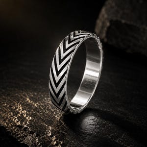 Puede incluir: Un anillo de banda de plata con un patrón de cheurón repetitivo grabado alrededor del exterior. El anillo tiene un acabado metálico pulido con áreas empotradas oscurecidas dentro de las ranuras en forma de V, creando un alto contraste.