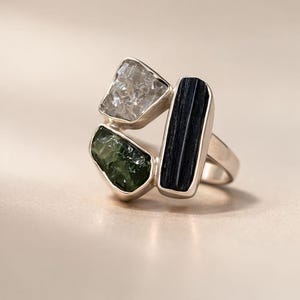 Può includere: Un anello in argento con tre distinte pietre grezze incastonate a grappolo. Le pietre includono un cristallo trasparente sfaccettato, una pietra grezza verde scuro e un lungo cristallo di tormalina nera orientato verticalmente su una fascia liscia.
