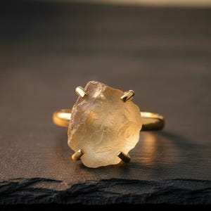 Puede incluir: Un anillo de oro con una gran piedra preciosa amarilla, cruda y translúcida, sujeta por cuatro garras metálicas. La piedra tiene una forma natural irregular con texturas internas visibles, montada sobre una banda de oro lisa y pulida.