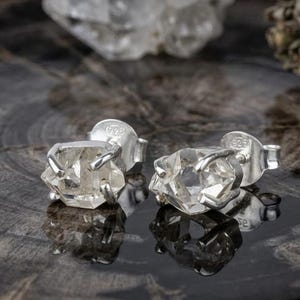 Peut inclure: Une paire de boucles d'oreilles à clous avec des cristaux de quartz brut transparents sertis dans des griffes en argent sterling. Chaque boucle porte le poinçon 925 sur le fermoir, mettant en valeur la texture naturelle et facettée des pierres.