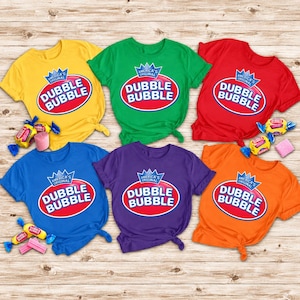 May include: Six short-sleeved t-shirts in yellow, green, red, blue, purple, and orange, each featuring the Dubble Bubble logo with a crown icon and the text America's Original. Several individually wrapped pieces of bubble gum are scattered around.