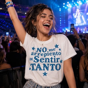 Op de afbeelding: Een wit T-shirt met korte mouwen en blauwe tekst die NO me arrepiento DE Sentir TANTO leest, versierd met blauwe sterren. De letters hebben een subtiel gespikkeld patroon en het T-shirt wordt gedragen met een blauwe spijkerbroek.