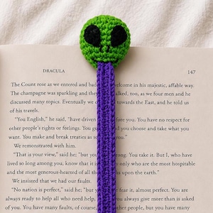 May include: A crocheted bookmark featuring a bright green alien head with large black oval eyes and a long, flat, textured purple tail extending downward, resting on the open pages of a book titled Dracula with visible printed text.