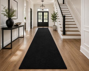 May include: A long, rectangular runner rug in a solid, deep black color. The rug features a low-pile, uniform texture and is positioned centrally on a light-toned wooden floor, extending from the foreground toward the back of the hallway.