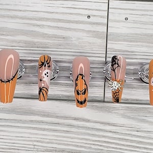 May include: Set of five Halloween-themed false nails. Each nail features a different design, including a jack-o'-lantern, a ghost, a spider web, and a crescent moon. The nails are painted in orange, black, and nude colors.