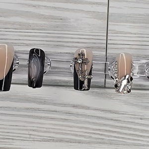 May include: A set of five artificial nails in various designs. One nail features a black and nude color block design, another has a swirling black and gray pattern, and a third has a cross charm. The other two nails have rhinestone and swirl designs.