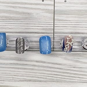 May include: A collection of four artificial nails with different designs. The nails are mounted on clear stands. Designs include blue with bubbles, silver with diagonal lines, blue with white lines, and a star design with rhinestones.