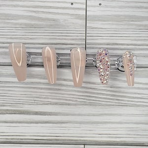 May include: Five long, coffin-shaped artificial nails. Three are a solid nude color, one is covered in iridescent rhinestones, and the last has a few rhinestones. The nails are displayed on a silver bar against a light wood-grain background.