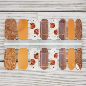 May include: Nail art strips featuring fall-themed designs. The strips include glitter, solid orange, and patterned designs with pumpkins, dots, and plaid. The color palette includes shades of orange, brown, and white, perfect for autumn.
