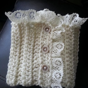 May include: Cream-colored, hand-crocheted item with a textured stitch pattern. Features a delicate white lace trim along the edges and is fastened with three small, light-brown buttons. The item appears to be a decorative accessory.
