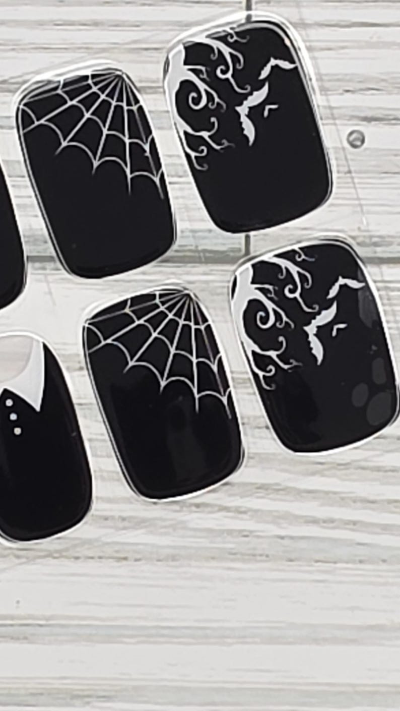 May include: Black nail art with white spiderwebs, bats, and decorative swirls. The nail designs are on a clear background, perfect for Halloween or gothic-themed looks. The nails are rectangular in shape.
