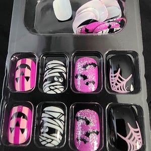 May include: A set of 10 Halloween-themed fake nails in a clear plastic case. The nails feature various designs, including pink and black stripes with a jack-o-lantern, a white mummy with black eyes, pink glitter with black bats, and black with a pink spiderweb.