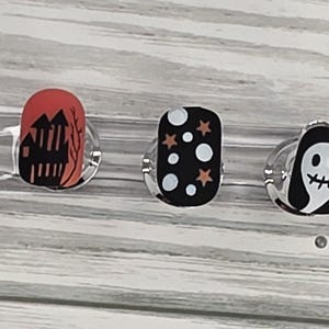 May include: A collection of Halloween-themed press-on nails. Designs include a jack-o'-lantern, haunted house, stars and dots, a ghost, and dripping orange and black. Each nail is oval-shaped and mounted on a clear display.